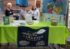 three women at a table outdoors with a Master Gardener sign and posters