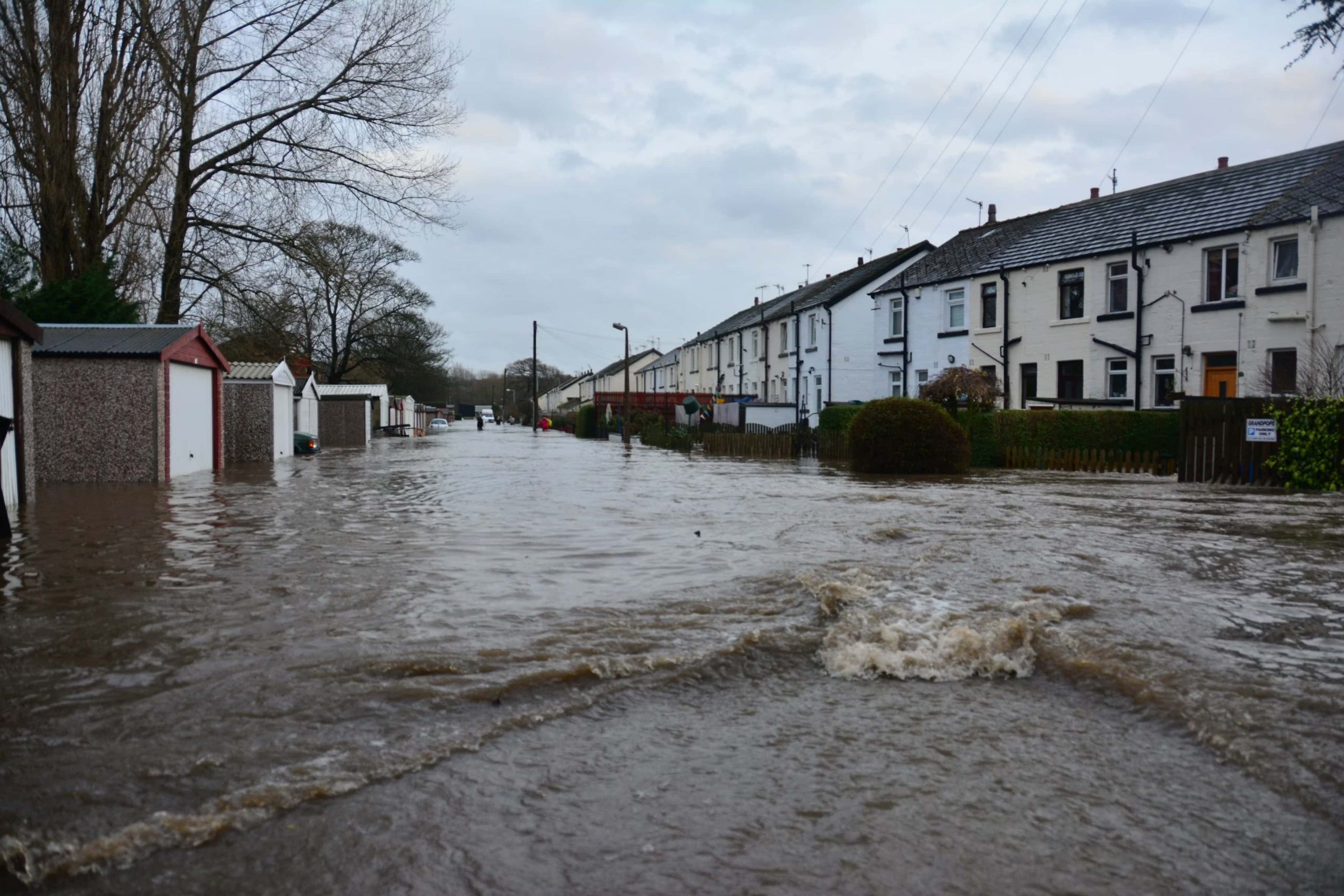 A flooded street with white houses on the right side, and small stone sheds on the left side.