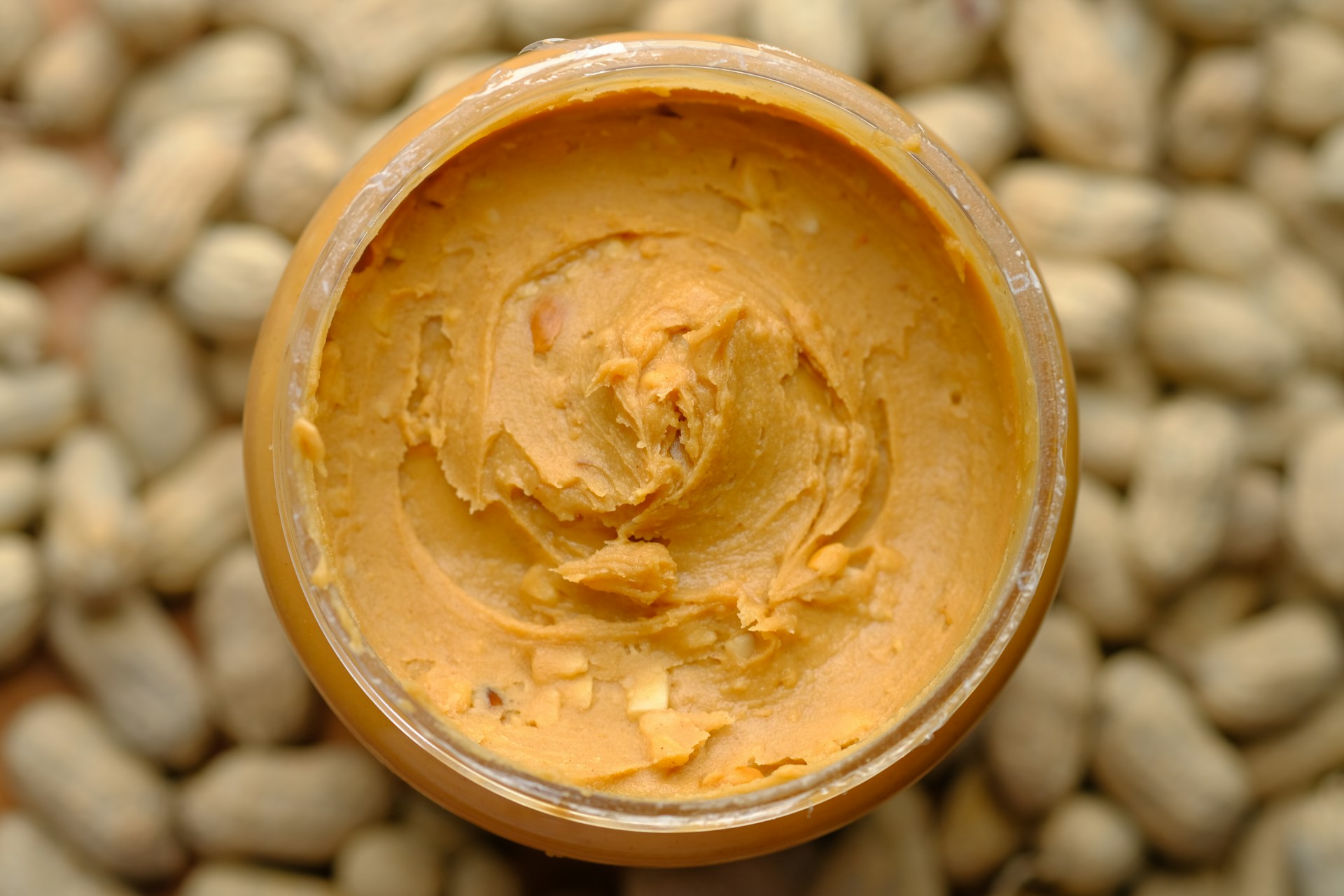 A photo loking into the top of a full jar of peanut buter,with peanuts on the table surounding the jar.
