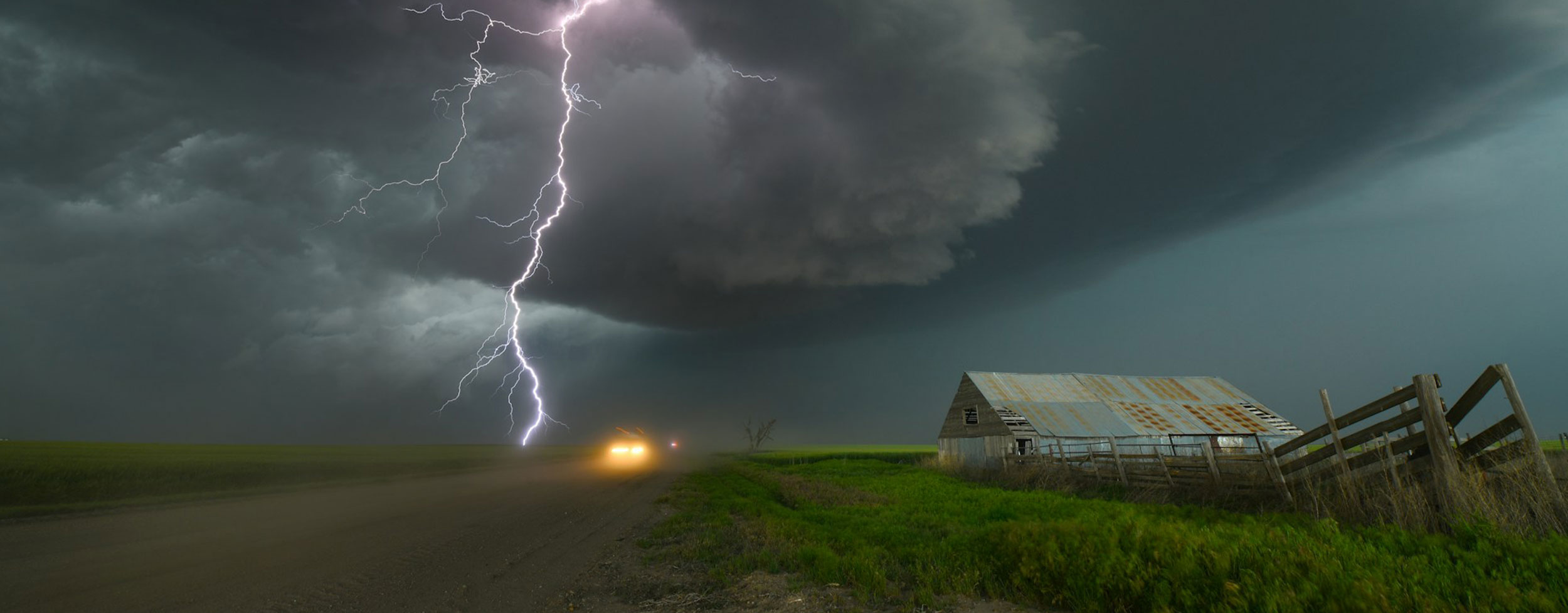 An image of a purple bolt of lightning striking the field on a farm with a wooden fence and metal barn on the right side of the image.