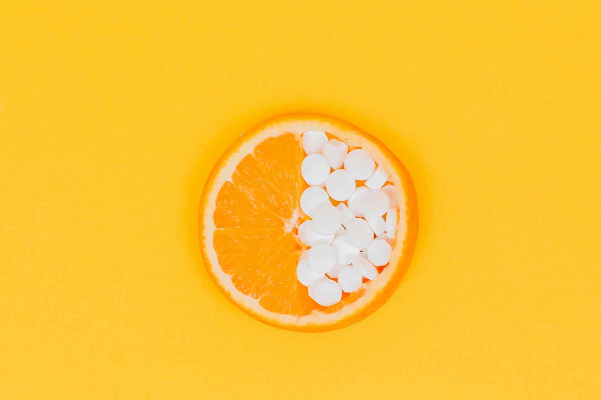 A slice of an orange with half of the circle filled with white tablet pills against a yellow background.