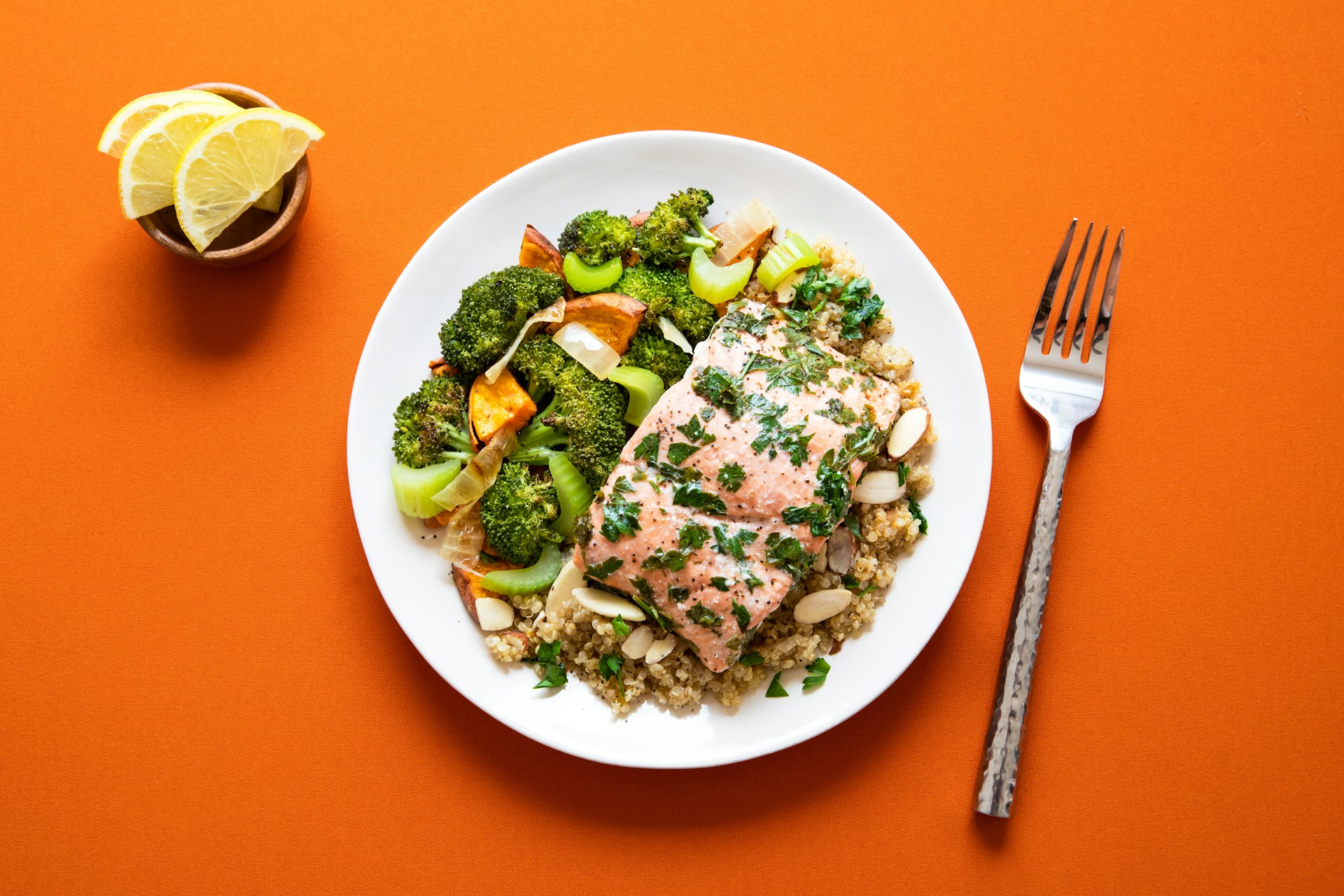 A photo looking down at a white plate of salmon, vegtables, and quinoa, with a silver fork on the right, and a wooden bowl with three slices of lemon on the left.