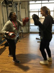 Two women participating in a boxing lesson.The woman on the left has red gloves, the one on the right has black training mits.