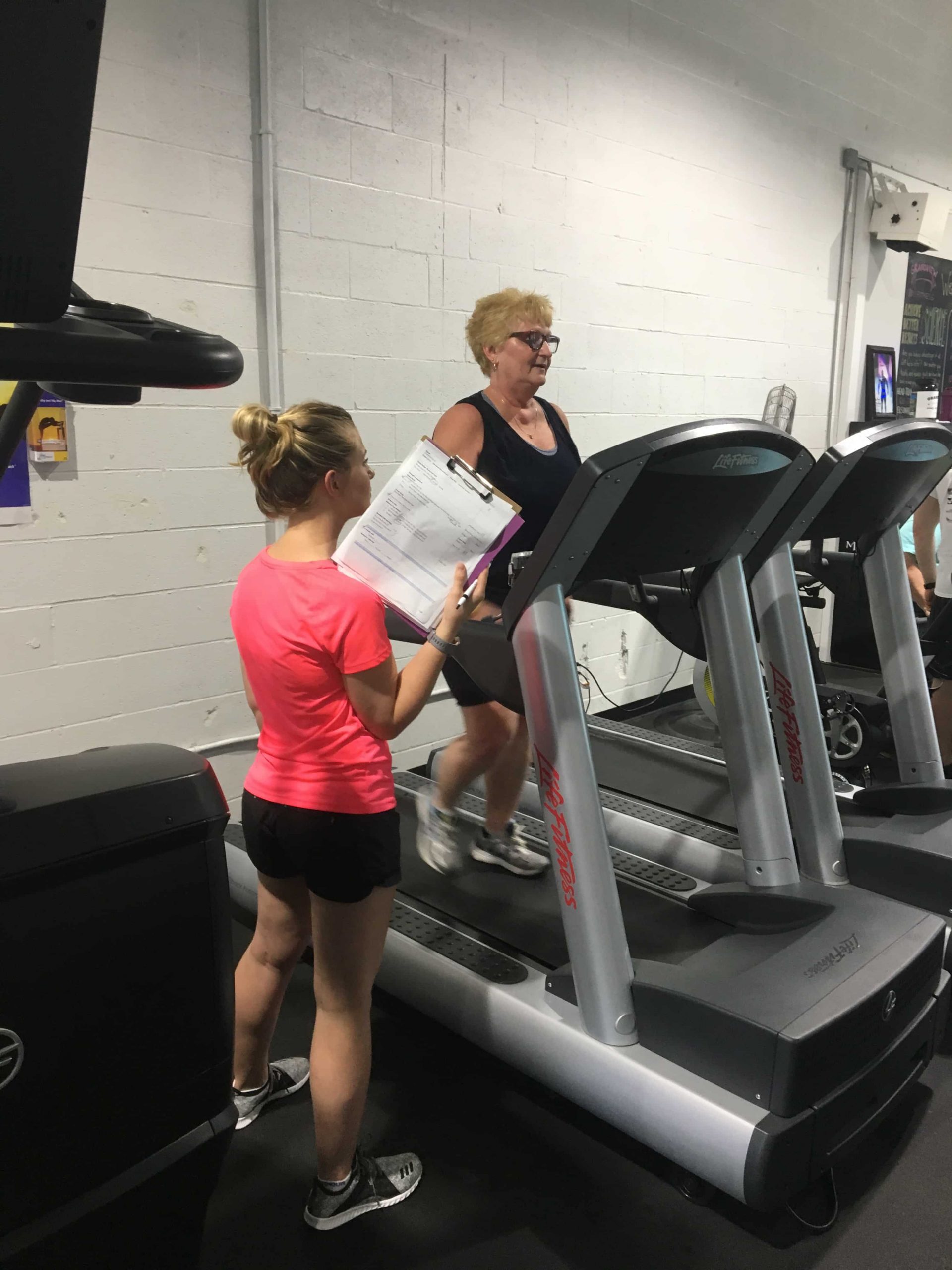 An older woman wearing black walks on a treadmill while a woman holding a clipboard stands next to her.
