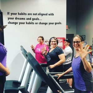 Three women smiling and waving at the camera while walking on treadmills.
