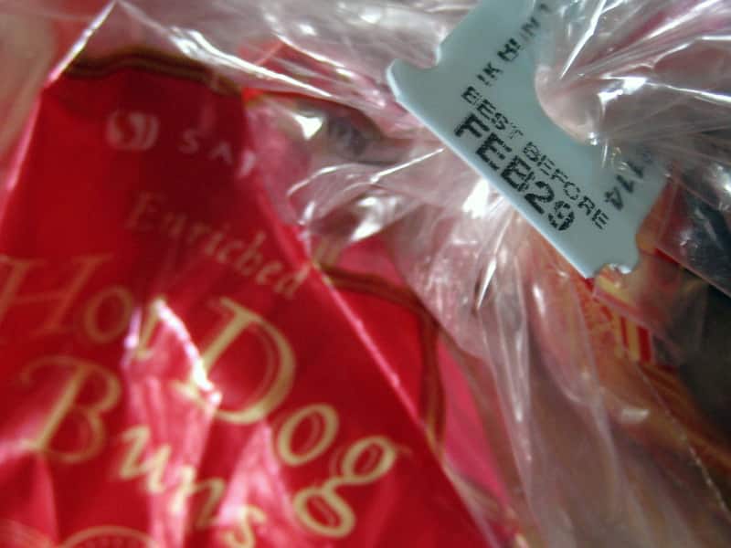 A close up image of a light blue bread expiration tag with the date "FEB 29" on a red bag of hot dog buns.