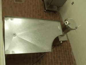A view of a silver automatic sausage stuffer on brick flooring against a white wall. The large metal table is attached to the machine.
