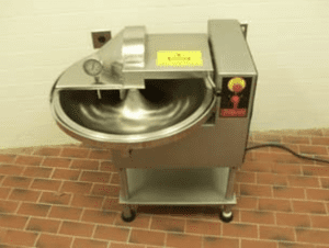 A silver bowl chopper in a room with brick flooring and white walls. The machine is connected to a black cord that goes out of view.