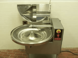 A silver bowl chopper in a room with brick flooring and white walls. The machine is connected to a black cord that goes out of view. The half lid of the chopper is open.