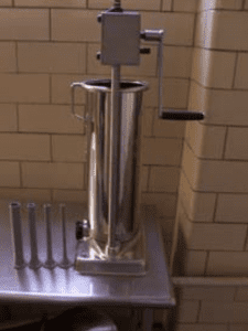 The side view of a silver handcrank sausage stuffer on a metal table.