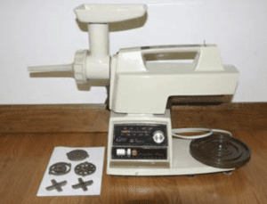 A white home sausage grinding/stuffing machine with a black square panel of controls on a wooden table. Machine attachments sit on a white towel on the table.