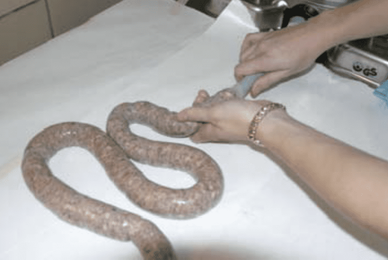 An individual wearing a gold bracelet on their left hand, utilising a machine to stuff meat into a natrual sausage casing.
