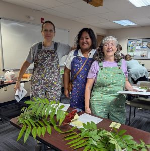 three women wearing gardening overalls and smiling with plants in the foreground