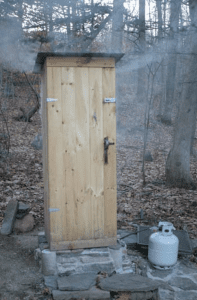 A closed smokehouse in a wooded area with trees and fallen leaves on the ground. Smoke is coming out of the structre which is connected to a smallgas tank on the ground.