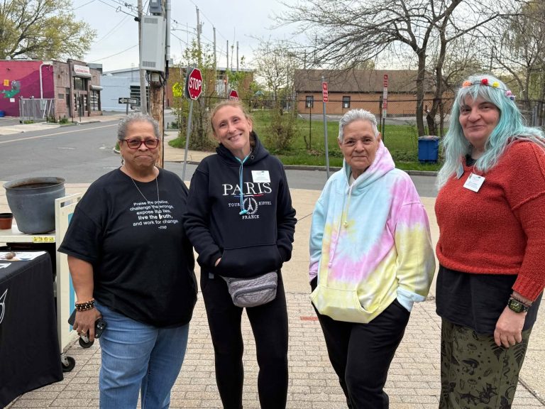 four women standing in an urban neighborhood with tables and garden supplies and educational materials