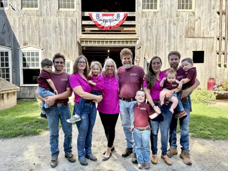 adults and children standing in front of a large barn with American flag bunting hanging on it and green grass alongside the driveway